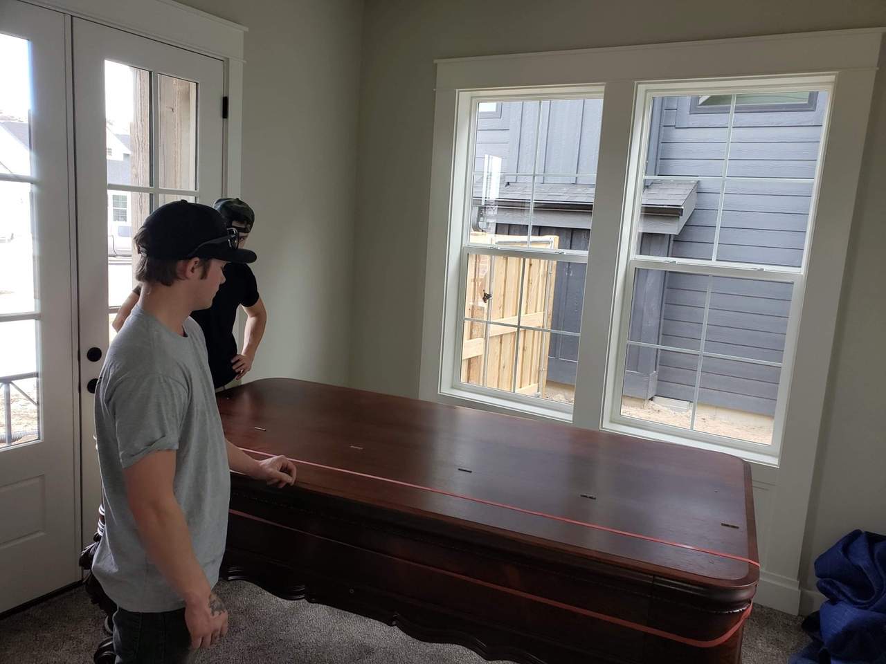 Two Delicate Touch movers lowering a long dark polished wood dining table into place in an empty, freshly painted room of a new build, a red lifting strap still running the length of the tabletop and a blue moving blanket on the carpet beside it